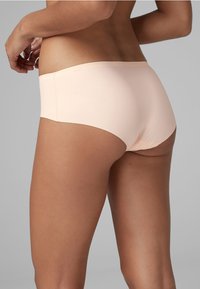 Vaalean persikanväriset hipster-alushousut, joissa on sileä pinta, matala vyötäröleikkaus ja saumattomat reunat mukavuutta varten. Takana näkyy minimalistinen design.