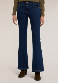 Jeans acampanados de denim azul con un lavado oscuro, que presentan bolsillos delanteros, pasadores para cinturón y costuras en contraste. Combinados con botas de tacón marrones.