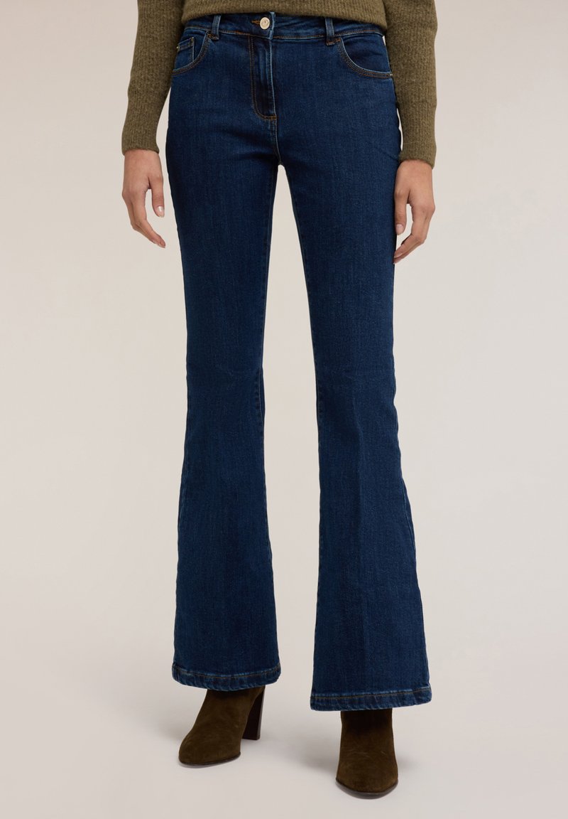 Jeans acampanados de denim azul con un lavado oscuro, que presentan bolsillos delanteros, pasadores para cinturón y costuras en contraste. Combinados con botas de tacón marrones.