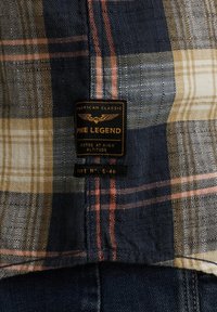 Chemise à carreaux dans des couleurs marine, beige et orange avec une texture tissée. Comprend une étiquette noire portant la mention "PME Legend" et des détails d'entretien.