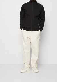 Veste zippée noire avec des manches raglan, associée à un pantalon crème fuselé et des baskets blanches, soulignant un look décontracté et contemporain.