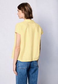 Blusa gialla a maniche corte con colletto, caratterizzata da una vestibilità ampia e un orlo leggermente curvo. Il tessuto sembra morbido con una texture liscia.