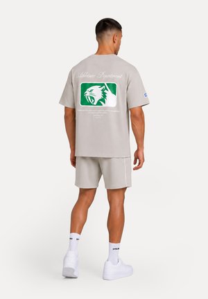 Mand iført beige shorts og t-shirt med en grøn og hvid brølende panter-logo på ryggen, hvide sokker og hvide sneakers, stående.