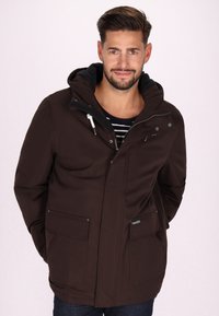 Giacca impermeabile marrone con cappuccio, dotata di due tasche frontali, zip argentate e dettagli con cordoncino. Indossata sopra una maglietta a righe.