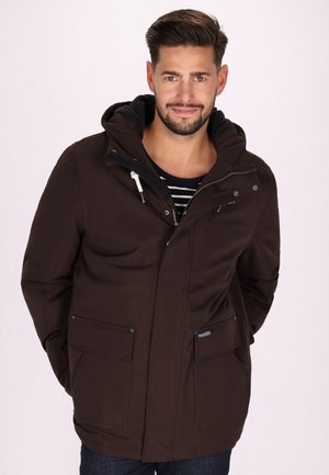 Bruine waterdichte jas met een capuchon, met twee voorzakken, zilveren ritsen en verstelbare accenten. Draagt over een gestreept shirt.
