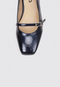 Escarpin en cuir verni marine avec un bout pointu et une bride de cheville, comportant une petite boucle en argent et une texture lisse et brillante.