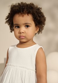 White sleeveless dress with ribbed texture and a gathered skirt, featuring a round neckline. Child with short, curly hair and a neutral background.