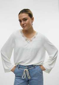Hvid langærmet bluse i tekstureret stof, med en blondekrave med blomstermønstre og en subtil V-form. Matchende med blå denim.