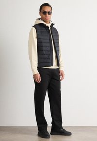 Μαύρη γιλέκο με φερμουάρ μπροστά, φορεμένη πάνω από μια ανοιχτό μπεζ φούτερ. Μαύρο παντελόνι και αθλητικά μαύρα sneakers ολοκληρώνουν την εμφάνιση.