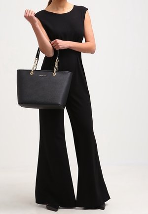 Femme en combinaison noire sans manches tenant un sac cabas en cuir noir avec des anses en chaîne dorée, debout devant un fond uni.