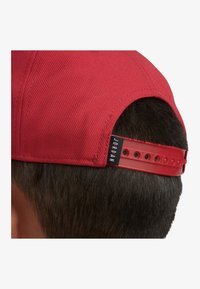 Sarkans cepure ar teksturētu virsmu, kurai ir plastmasas regulējama josta un melns birka ar uzrakstu "JORDAN" aizmugurē.