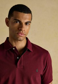 Burgundia polo särk musta kraega, kahe valge nupuga ja väikese rohelise logoga rinnal. Kangas tundub sile ja tekstureeritud.