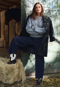 Szary sweter w splot warkoczowy, czarny kwiatowy płaszcz, granatowe spodnie w prążki oraz czarne botki z białymi akcentami. W pierwszym planie balot siana.