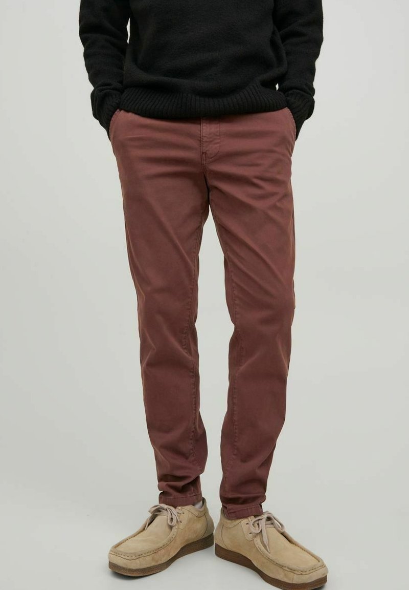 Personne portant un pantalon couleur rouille, un pull noir, des chaussures décontractées beige avec lacets, debout les mains dans les poches devant un fond uni.