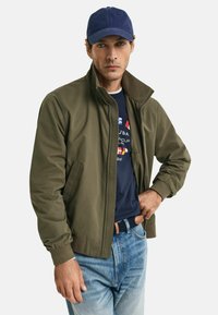 Olive green bomber jacket with a high collar, paired with a navy cap, navy t-shirt, and light blue jeans. Features ribbed cuffs and side pockets.