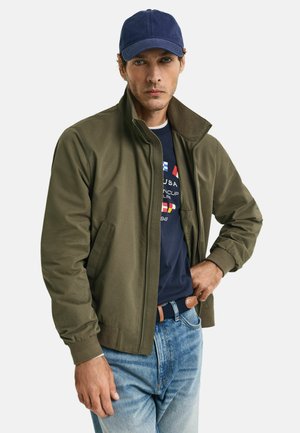 Olive green bomber jacket with a high collar, paired with a navy cap, navy t-shirt, and light blue jeans. Features ribbed cuffs and side pockets.