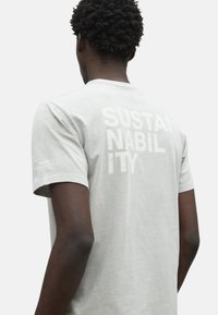 Camiseta de algodón de color gris claro, con el texto en bloque "SUSTAINABILITY" impreso en la parte posterior en un tono ligeramente más oscuro. Mangas cortas.