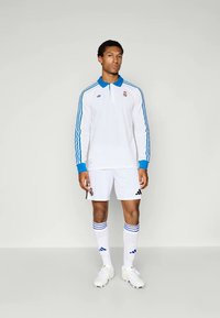 Hvit polo med lange ermer og blå krage og striper, hvite shorts, og matchende hvite sokker med blå detaljer, brukt med hvite joggesko.