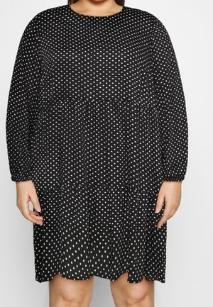 Czarna sukienka z długim rękawem w białe kropki, luźnego kroju, o długości do połowy uda, noszona przez osobę ze swobodnie opuszczonymi rękami.