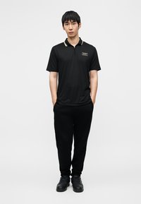 Μαύρο πόλο μπλουζάκι με φερμουάρ στον γιακά, χρυσές λεπτομέρειες και λογότυπο· συνδυασμένο με μαύρο παντελόνι φόρμας και μαύρα αθλητικά παπούτσια. Απλός, αθλητικός σχεδιασμός.