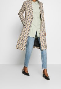 Abrigo trench de doble botonadura en patrón de cuadros amarillos y negros, combinado con una camisa verde claro y jeans azules. Zapatos de tacón negros.