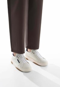Sneaker bianche perforate con suole in gomma marrone chiaro, accentuate da dettagli grigi e blu navy, abbinate a pantaloni marroni larghi.