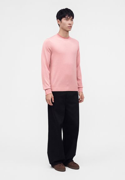 Jeune homme debout portant un pull rose à manches longues, un pantalon large noir et des chaussures marron sur un fond clair uni.