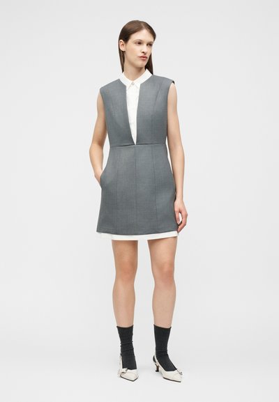 Γυναίκα με γκρι φόρεμα χωρίς μανίκια πάνω από λευκό πουκάμισο, συνδυασμένο με σκούρες κάλτσες και λευκά ψηλά τακούνια, στέκεται με το ένα χέρι στην τσέπη.