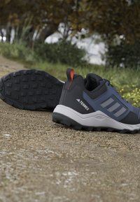 Chaussures de randonnée Adidas Terrex noires et bleues avec semelles robustes reposant sur un chemin de gravier avec de la verdure en arrière-plan.