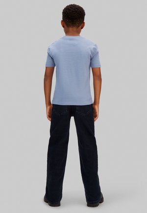 Παιδί που στέκεται με την πλάτη στο θεατή, φορώντας ανοιχτό μπλε κοντομάνικο πουκάμισο, σκούρο παντελόνι και μαύρα παπούτσια, μπροστά σε απλό φόντο.