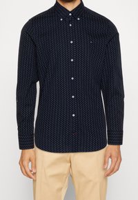 Man in een navyblauw overhemd met witte stippen en beige broek, staand met de armen ontspannen langs het lichaam.