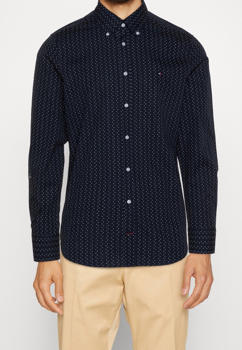 Man in een navyblauw overhemd met witte stippen en beige broek, staand met de armen ontspannen langs het lichaam.