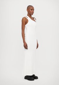 White, ribbed maxi dress with a fitted bodice, featuring asymmetric straps and side slits, paired with black chunky boots.