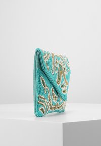 Clutch in tessuto turchese con ricami di perline dorate e bianche in motivi a spirale, posata su un pedestal bianco contro uno sfondo grigio.