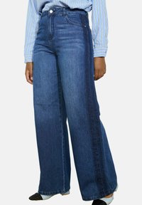 Wijdvallende jeans in medium blauwe denim met een donkerdere zijpaneel. Beschikt over vijf zakken, riemlussen en een hoge taille.