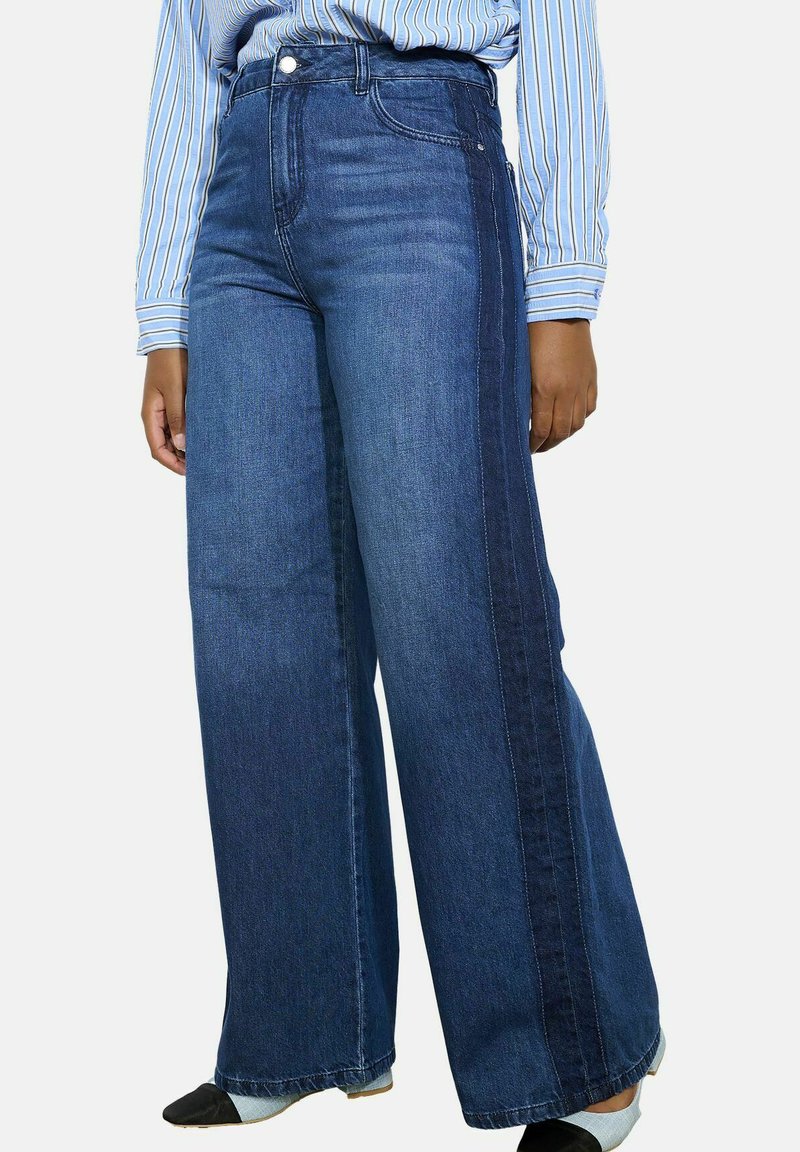 Wijdvallende jeans in medium blauwe denim met een donkerdere zijpaneel. Beschikt over vijf zakken, riemlussen en een hoge taille.