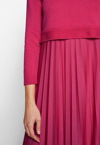 Main d'une femme reposant à côté d'une jupe plissée fuchsia et d'un pull en maille à manches longues assorti sur un fond blanc.