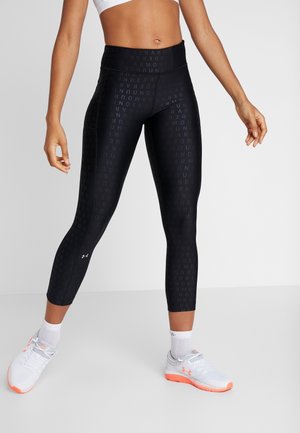 Vrouw die zwarte legging met patroon van Under Armour draagt, witte sportsokken en witte hardloopschoenen met oranje zolen, staande met de voeten uit elkaar.