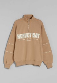 Sweatshirt zippé marron au col avec une coupe décontractée, orné d'un texte imprimé blanc "HERVEY BAY QUEENSLAND Australie" et d'accents blancs sur les manches.