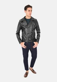 Giacca da biker in pelle nera con cerniere e bottoni a pressione argentati, caratterizzata da una vita aderente e una tasca a pattina. Indossata sopra un maglione scuro e jeans blu.