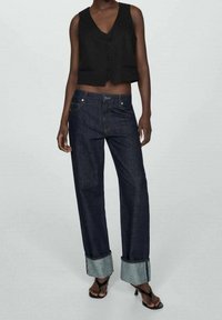 Vest cropped nero con bottoni abbinato a jeans in denim scuro a vita alta, con risvolti di un blu chiaro e dettagli di cucitura sottili.