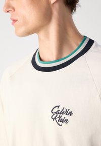 Krémové tričko s námořnickým a tyrkysovým pruhovaným výstřihem do kulata, se tmavým vyšitým logem "Calvin Klein" na prsou.