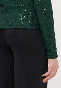 Haut en sequins verts à manches longues, porté sur un pantalon noir à côtes. Le haut présente des sequins brillants sur une surface texturée.