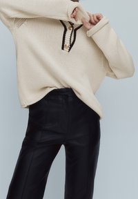 Personne portant un pull en maille beige ample avec des boutons dorés et des bordures noires, assorti à un pantalon en cuir noir taille haute, sur un fond gris.