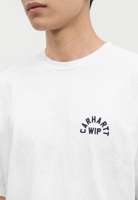 Крупен план на човек, който носи бяла тениска с малко черно лого "CARHARTT WIP" на лявата гърда.