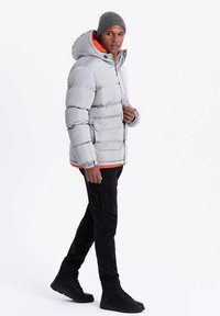 Veste en duvet grise avec capuche, dotée d'une doublure orange, de poches avant et de manches côtelées, associée à un pantalon cargo noir et des bottes noires.