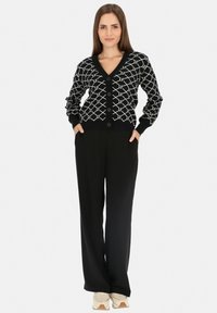 Czarny kardigan z białym geometrycznym wzorem, dekolt w serek, z przodu guziki i długie rękawy; połączony z czarnymi spodniami o szerokich nogawkach i jasnymi sneakersami.