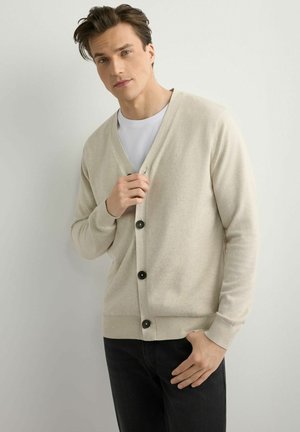 Mężczyzna ubrany w beżowy sweter na guziki, na biały shirt i czarne spodnie, poprawiający sweter lewą ręką, stojący na neutralnym tle.