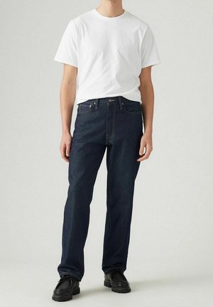 Mężczyzna ubrany w prosty biały t-shirt, ciemnoniebieskie dżinsy i czarne buty stojący na jasnoszarym tle.