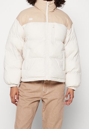 Person wearing a beige and white padded jacket with a zip front, standing with hands relaxed at sides against a plain white background.